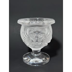 Könnte beinhalten: Ein Votivkerzenhalter aus klarem, satiniertem Glas mit einem skulpturalen Löwenkopf-Relief auf der Vorderseite. Das Gefäß verfügt über einen breiten, flachen Rand, einen abgerundeten Körper und einen stabilen runden Fuß aus durchscheinendem Glas.