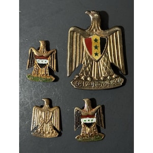 May include: Four vintage metal badges featuring eagle designs. The badges are gold-toned with intricate details, including wings, shields, and Arabic script. The shields display flags and stars. The badges vary in size and shape, with a dark background.