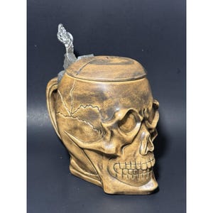 以下が含まれることがあります： 人間の頭蓋骨の形をしたセラミック製のビールジョッキ。ジョッキはタン色で、濃い茶色の陰影があり、金属製のヒンジ付きの蓋とハンドルが付いています。頭蓋骨には詳細な顔の特徴と表面のひび割れがあります。
