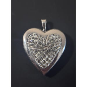 Puede incluir: Un colgante en forma de corazón de tono plateado con una incrustación central llena de brillantes piedras de estrás transparentes. Una figura de ángel de plata pulida con alas extendidas se sitúa sobre el centro de la superficie del corazón.