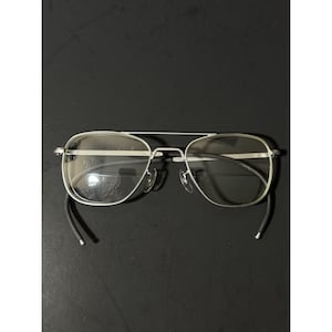 May include: Silver-framed aviator eyeglasses with clear lenses. The glasses have a double bridge and black temple tips. The lenses are oval-shaped and the frame is a shiny silver color. The glasses are set against a black background.