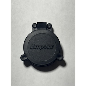 Op de afbeelding: Een zwarte Aimpoint lensdop. De dop is rond met de merknaam in het midden. De dop heeft twee kleine cirkelvormige uitsteeksels aan de bovenkant en twee aan de zijkanten.