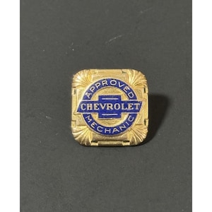 Puede incluir: Un pin de metal dorado vintage con el texto "APPROVED CHEVROLET MECHANIC" en azul y blanco. El pin es cuadrado con un borde decorativo.