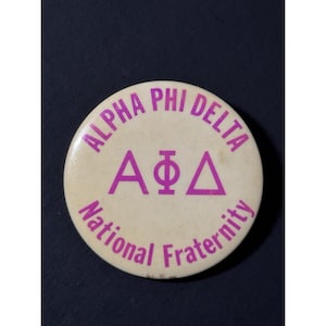 May include: A circular, cream-coloured button with the words "ALPHA PHI DELTA" in a purple, curved font. The centre displays the Greek letters A, Φ, and Δ in purple. The words "National Fraternity" are also in purple.