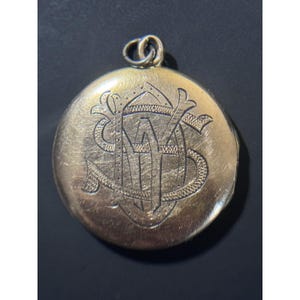 May include: A round, gold-toned metal locket featuring an intricate, engraved monogram on its front surface. The design consists of overlapping, stylized letters with textured detailing. A small, circular metal jump ring is attached to the top.