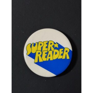 May include: A round, white button with the words "SUPER READER" in yellow, outlined in blue. A blue diamond shape is behind the text. The button is a fun accessory for book lovers.