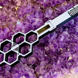 Könnte beinhalten: Ein weißes und schwarzes Werkzeug mit Wabenmuster und dem Wort "ember" auf der Seite. Das Werkzeug hat einen goldfarbenen Akzent und liegt vor einem Hintergrund aus violetten Amethystkristallen.