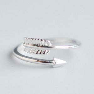 May include: A silver ring with a delicate arrow design. The arrow is wrapped around the band of the ring, with the arrowhead pointing to the right.