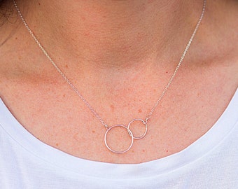 Pode incluir: Um delicado colar de prata com dois círculos abertos interligados. O pingente pende de uma corrente fina, criando um design minimalista e elegante. O colar é mostrado sobre uma blusa branca.