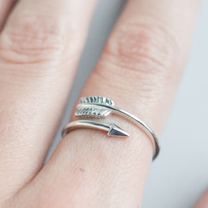 May include: A silver ring with a delicate arrow design. The arrow is wrapped with two small feathers.