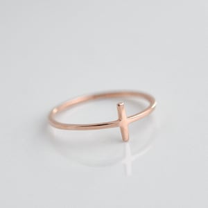 May include: A simple gold-toned ring with a thin cross design. The ring is on a white background.