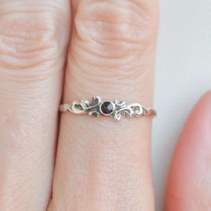 Puede incluir: Un anillo de plata con un diseño ornamentado y una pequeña gema negra en el centro. El anillo tiene un delicado patrón de estilo filigrana con detalles florales y de volutas. El anillo se muestra en un dedo.