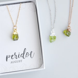 May include: A white box with a gold necklace featuring a raw peridot stone pendant. The box has the text "peridot AUGUST" printed on it. Two other necklaces with raw peridot stone pendants are visible in the background.
