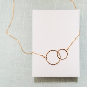 May include: A gold necklace with two interlocking circles pendant. The necklace is on a white background.