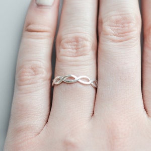 May include: A silver ring with a braided design. The ring is worn on a person's finger.