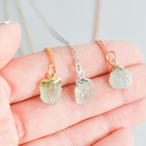May include: Three delicate necklaces with raw crystal pendants. Each necklace features a different metal chain: gold, silver, and rose gold. The crystal pendants are a light green color.