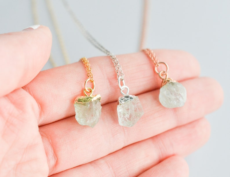 May include: Three delicate necklaces with raw crystal pendants. Each necklace features a different metal chain: gold, silver, and rose gold. The crystal pendants are a light green color.