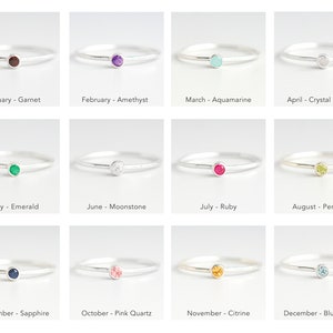 May include: Twelve silver rings with a small gemstone in the center. Each ring represents a different month of the year and features a corresponding birthstone. The birthstones are: Garnet, Amethyst, Aquamarine, Crystal Quartz, Emerald, Moonstone, Ruby, Peridot, Sapphire, Pink Quartz, Citrine, and Blue Topaz.