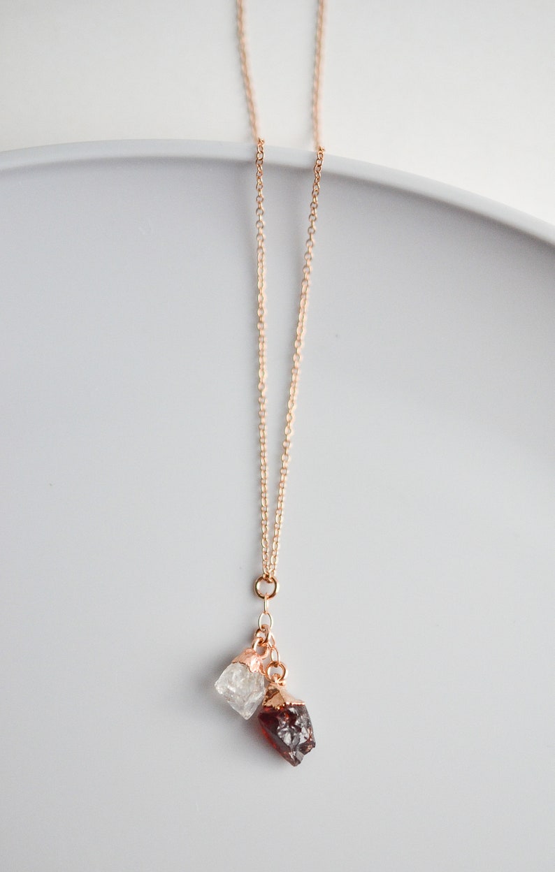 Puede incluir: Un collar de cadena dorada con dos colgantes de piedras preciosas en bruto. Un colgante es un cristal de cuarzo transparente y el otro es un granate rojo oscuro.