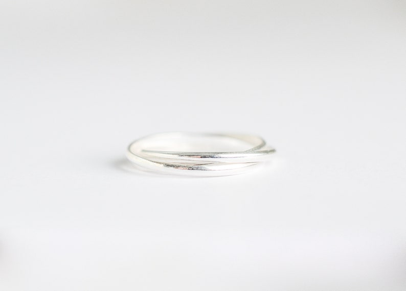 Pu&ograve; includere: Un anello d'argento con un design a tripla fascia. L'anello ha una finitura liscia e lucida e uno stile semplice ed elegante. Le fasce si intrecciano per creare un look unico e moderno. L'anello &egrave; su sfondo bianco.
