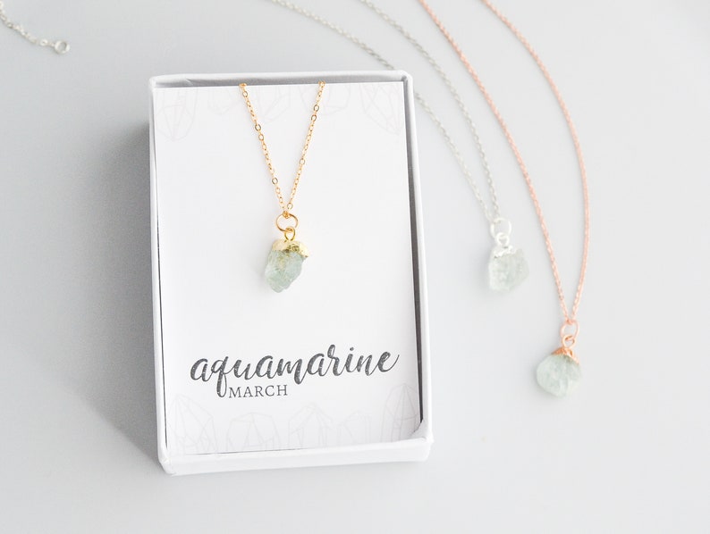 May include: Three necklaces with aquamarine crystal pendants in gold, silver, and rose gold settings. The necklaces are displayed in white boxes with the text "aquamarine MARCH" on the box.