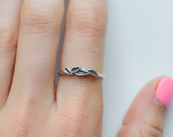 Op de afbeelding: Een zilveren ring met een delicaat bladerontwerp. De ring heeft een dunne band en een cluster van drie bladeren. De bladeren hebben gedetailleerde nerven, wat textuur toevoegt. De ring wordt getoond op een vinger met een roze nagel.