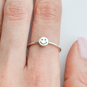 May include: A silver ring with a small smiley face charm. The ring is worn on a person's finger.