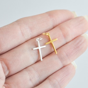 Sterling silver and gold cross charms held in a hand against a white background, showcasing their small size and delicate minimalist design.