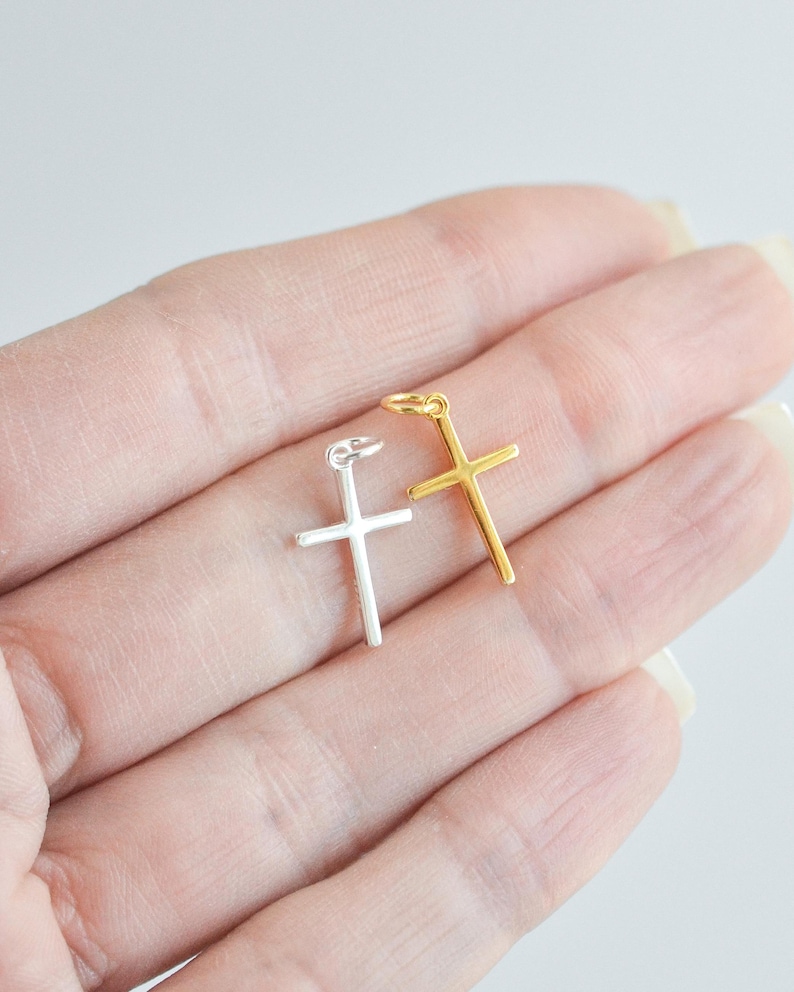 Sterling silver and gold cross charms held in a hand against a white background, showcasing their small size and delicate minimalist design.
