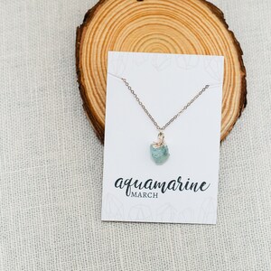 May include: A delicate gold chain necklace with a light blue aquamarine gemstone pendant. The necklace is displayed on a white card with the text "aquamarine MARCH".
