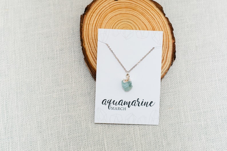 May include: A delicate gold chain necklace with a light blue aquamarine gemstone pendant. The necklace is displayed on a white card with the text "aquamarine MARCH".