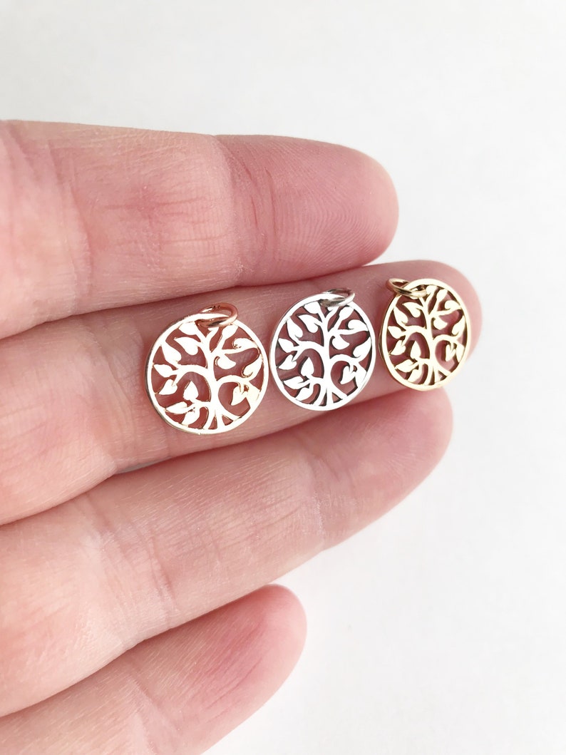 May include: Three small, round pendants with a tree of life design. One is gold-colored, one is silver-colored, and the other is gold-colored. The pendants are held in a hand against a white background. The tree design is cut out of the metal.