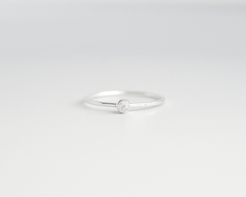 May include: A delicate silver ring with a small, round, clear gemstone set in the center. The ring band is thin and polished, creating a minimalist and elegant design. The ring is set against a plain white background.