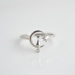May include: A silver ring with a cat sitting on a crescent moon and a star to the side.