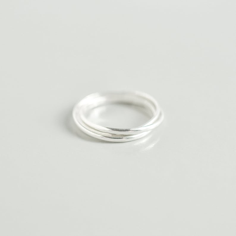 Pu&ograve; includere: Un anello d'argento con tre fasce intrecciate. Le fasce sono lisce e lucenti, riflettendo la luce. L'anello &egrave; posto su uno sfondo grigio chiaro, evidenziando la sua semplice eleganza. Un gioiello minimalista.