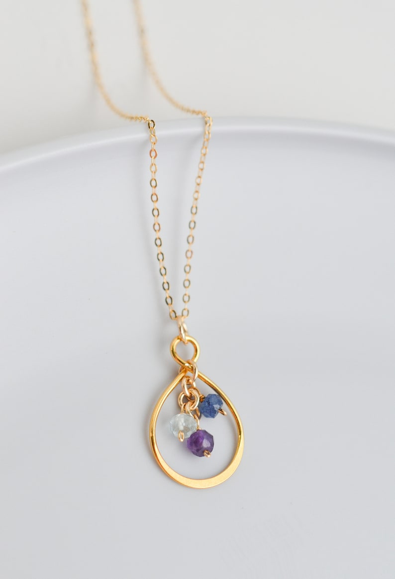May include: A gold chain necklace with a teardrop-shaped pendant. The pendant has three small gemstones: a light blue aquamarine, a dark blue sapphire, and a purple amethyst.