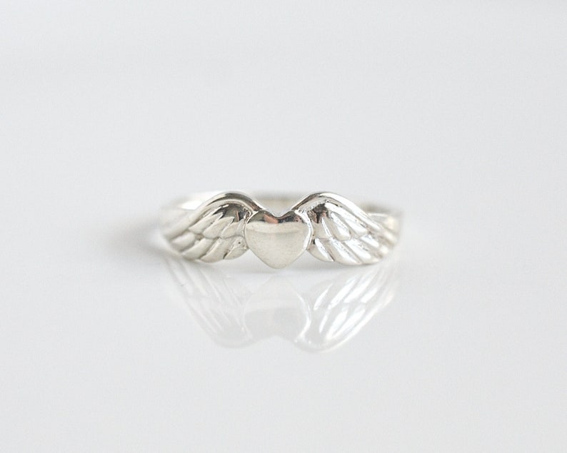 May include: A silver ring with a heart-shaped design and wings on either side.