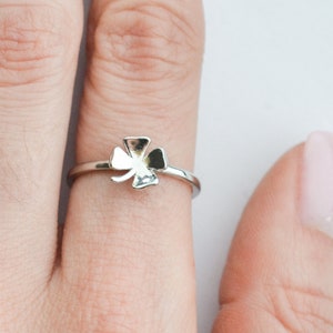 May include: A silver-colored ring featuring a four-leaf clover design. The ring is shown on a finger, with the clover charm centered on the band. The ring's band is thin and delicate, and the clover has a detailed, textured appearance.