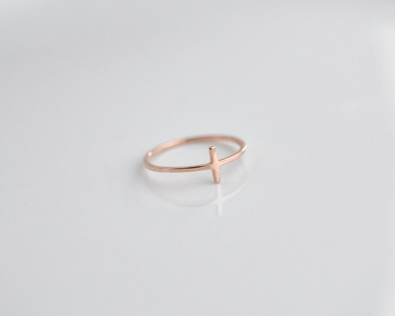 May include: A simple gold-toned ring with a thin cross design. The ring is on a white background.