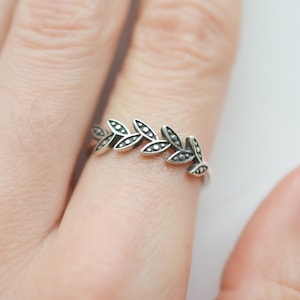 May include: A silver ring featuring a delicate design of leaves. The leaves are arranged in a row and have a slightly textured surface.