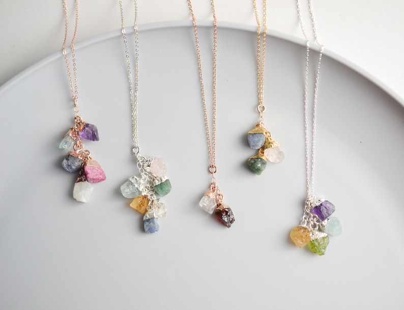 Puede incluir: Cinco delicados collares con dijes de piedras preciosas en bruto. Cada collar presenta un racimo de piedras preciosas de diferentes colores, incluyendo amatista, aguamarina, cuarzo rosa, aventurina verde y citrino.