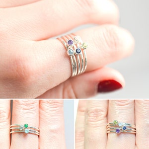 May include: Set of four silver stacking rings with small gemstones. The rings feature a light blue, green, purple, and yellow gemstone.