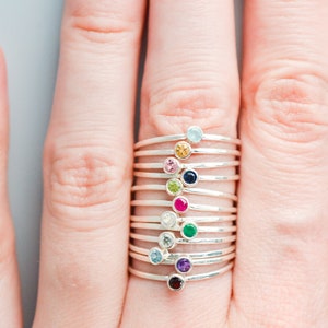 May include: A stack of silver rings with different colored gemstones. The gemstones are aquamarine, yellow, pink, blue, green, red, white, emerald, amethyst, and garnet.