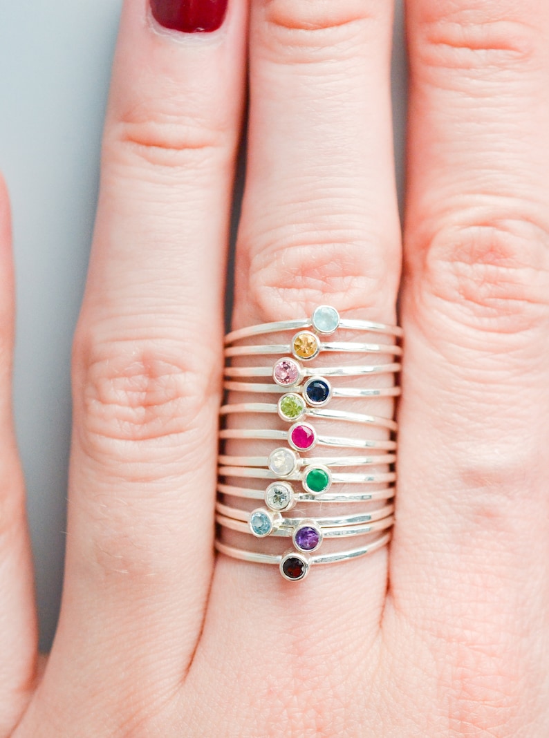 May include: A stack of silver rings with different colored gemstones. The gemstones are aquamarine, yellow, pink, blue, green, red, white, emerald, amethyst, and garnet.