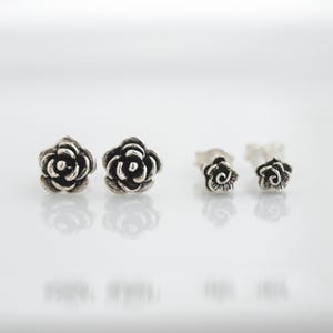 May include: Two pairs of silver rose stud earrings. The larger pair features detailed petals, while the smaller pair has a simplified design. The earrings are displayed on a white surface, reflecting the light.