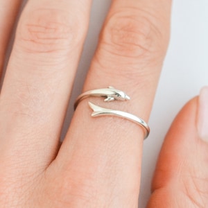 May include: A silver ring with a dolphin design. The dolphin is jumping out of the water and the ring is wrapped around a finger.