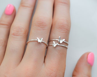 Op de afbeelding: Een close-up van een hand met drie delicate zilveren ringen met vlinderontwerpen. De ringen zijn gestapeld op de middelvinger.