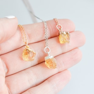 May include: Three necklaces with raw citrine crystal pendants. One necklace has a gold chain, one has a silver chain, and the third has a rose gold chain. The citrine crystals are a light orange color and have a rough, natural texture.