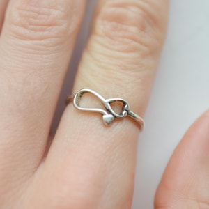 May include: A silver ring featuring an infinity symbol design with a small heart detail. The ring is worn on a finger, showcasing its delicate and minimalist style. The ring's design is simple yet elegant, suitable for everyday wear.
