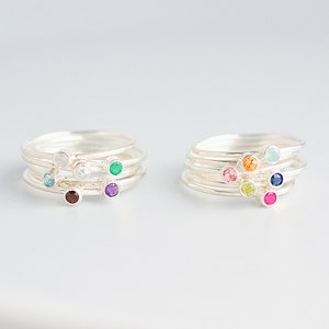 May include: Set of five silver stacking rings with colorful gemstones. The rings feature a variety of colors, including red, green, blue, purple, and white.